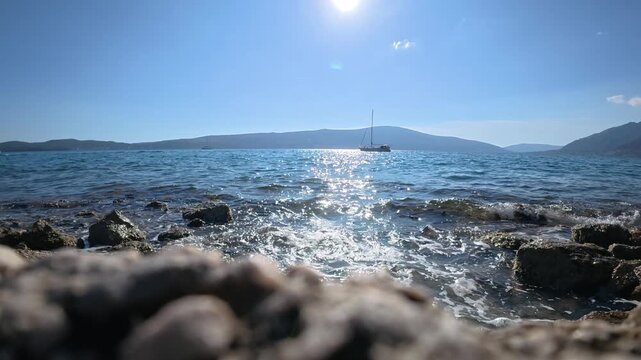 Slow motion sea waves splashing on rocky shore with yacht in distance