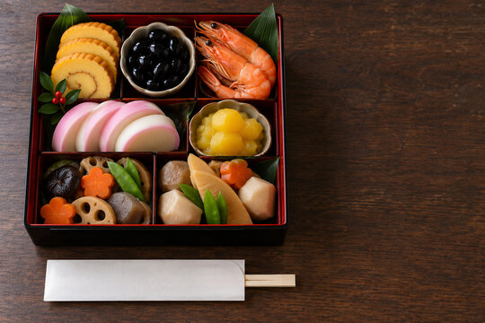 Osechi ryori in a black and red lacquer jubako box with rolled omelet, black soybeans, shrimp, kamaboko, kinton and simmered vegetables on a dark wooden table, top view with copy space