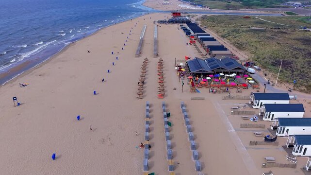 High-angle aerial drone clip of wide sandy beach shoreline, North Sea waves, beach cabins, resort pavilion, and coastal dunes in Katwijk aan Zee, South Holland, Netherlands on bright summer day