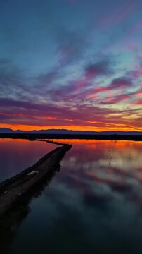 Cinematic vertical 9:16 aerial video flying over the calm waters alongside a dirt path at Kalochori Lagoon in Thessaloniki, Greece. The scene features a breathtaking sunset with vibrant purple, magent