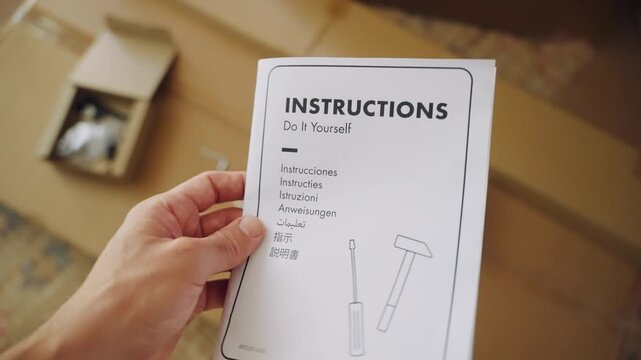 First person view of a man reading a multilingual instruction manual before assembling new flat pack furniture, with tools and components lying on the cardboard box in the background at home
