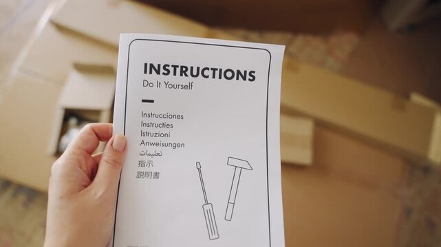 First person perspective of a person holding a DIY instruction manual before assembling new furniture, with cardboard boxes and tools visible in the blurred background of the living room