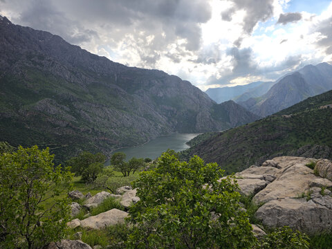 Lush green trees and boulders in the foreground, with a river in a deep valley, steep rocky mountains, scattered trees on slopes and dramatic clouds.
📍 Hawraman, Sarvabad, Kurdistan 