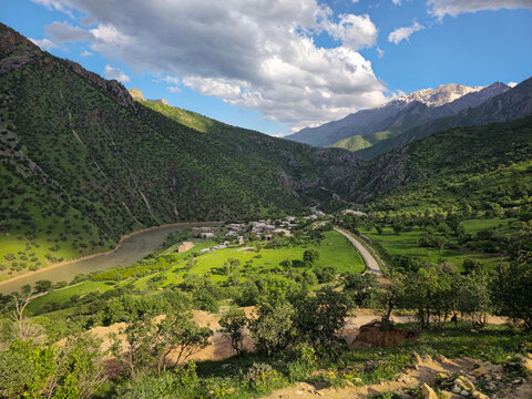 Mountain village nestled in a lush sunlit valley beside a winding river, with road, green meadows, yellow wildflowers, tree-covered mountains, fluffy clouds and distant snow-dusted peaks.
📍Zom
