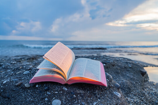 Open Bible Book with Fluttering Pages on Rocky Beach at Sunset