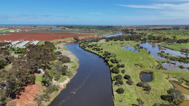 Aerial drone view following the Werribee River in Werribee South, Melbourne, Victoria, Australia, with flooded river water winding between eroded banks, eucalyptus bushland, and ploughed farmland.