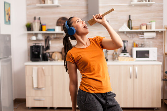 Energetic woman in headphones uses a wooden spoon as a pretend microphone while singing to music during a playful moment in her home kitchen.