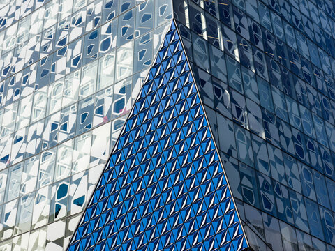 Close-up of a contemporary building corner where a triangulated blue mosaic-tiled spire rises between two glass curtain wall facades patterned with faceted geometric motifs.