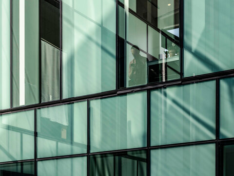 Exterior view of a business office building's glass curtain wall, showing a man sitting inside at a desk looking at his phone.