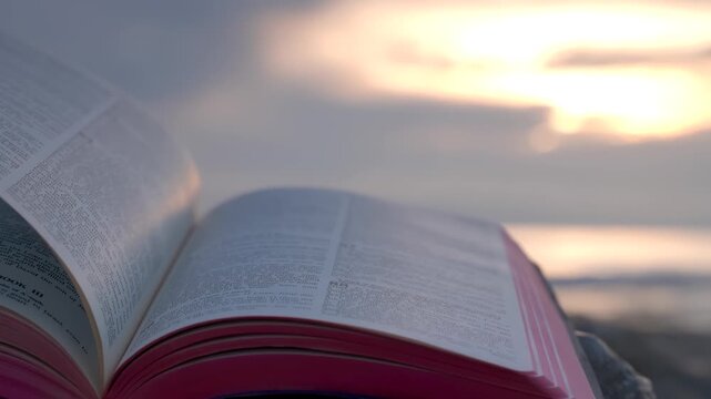 Open Bible Book with Fluttering Pages on Rocky Beach at Sunset