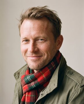 Confident mature man in red plaid scarf and olive jacket with welcoming expression, neutral backdrop