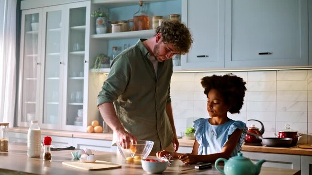 Father cracking eggs bowl with daughter. Multiracial family baking home kitchen.