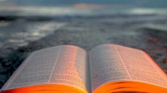 Open Bible Book with Fluttering Pages on Rocky Beach at Sunset