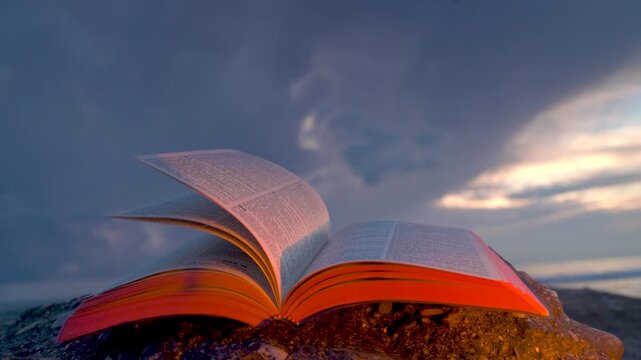 Open Bible Book with Fluttering Pages on Rocky Beach at Sunset