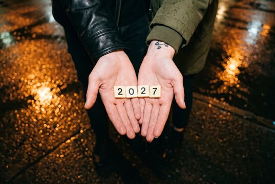 Open palms presenting 2027 number tiles on city street with golden lights, symbol of the future