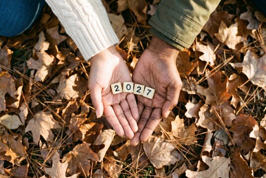 Open hands side by side presenting 2027 number tiles on fallen leaves, concept of renewal