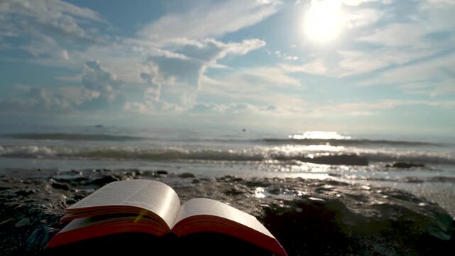 Open Bible Book with Fluttering Pages on Rocky Beach at Sunset