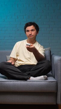 Vertical video man relaxing on a sofa in his living room, feeling bored and unengaged while searching for something interesting to watch on television using a remote control