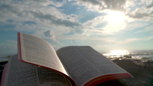 Open Bible Book with Fluttering Pages on Rocky Beach at Sunset