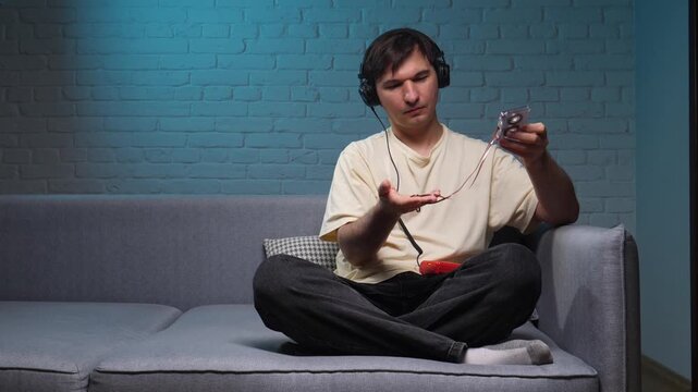 Young man wearing headphones looking at a broken cassette tape with tangled magnetic tape, experiencing frustration and nostalgia for retro analog media on a couch