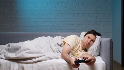 Man reclines in bed under a blanket holding a game controller, gaming alone at night for relaxed leisure and casual entertainment in a cozy home bedroom setting © Aliaksandr