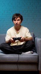 Young adult man engrossed in playing a video game, holding a wired controller while sitting cross legged on a gray couch in a living room with a blue brick wall © Aliaksandr