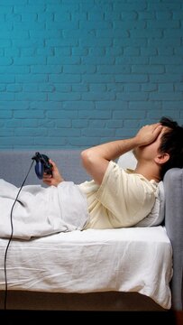 Young man lying on a bed covering his face in disappointment and sadness after losing a video game while holding a controller, expressing feelings of failure and anger