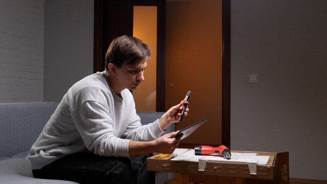 Young man concentrating on digital instructions on a tablet, holding pliers while sitting on a couch, diligently assembling furniture from a flat pack box at home