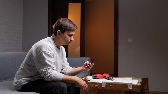 Young man sitting on a sofa and looking at a smartphone screen, searching for diy furniture assembly instructions with tools and flat pack parts on a nearby table