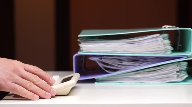 Hand calculating numbers on a desktop calculator, surrounded by stacked binders filled with financial documents and paperwork, symbolizing detailed business accounting and tax preparation