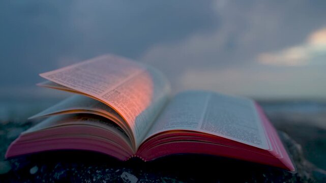 Open Bible Book with Fluttering Pages on Rocky Beach at Sunset