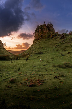 Sunset paints sky with warm orange and purple hues. Rocky cliff rises above lush green meadow. Two small figures stand atop cliff, silhouetted against fading light. Grasses sway gently in breeze acros