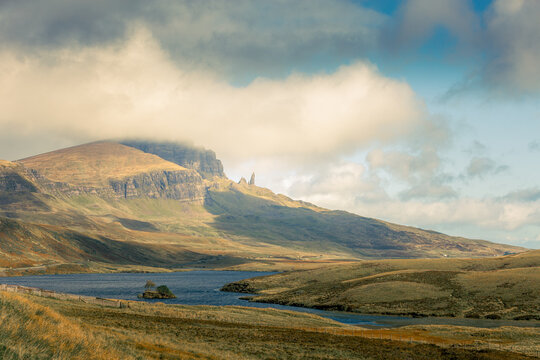 Sunlight breaks through clouds over misty mountain peak. Calm lake reflects sky and rugged terrain. Grasslands stretch beside water, peaceful and serene. No people present in this tranquil Scottish hi