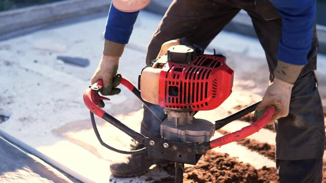 Worker operates red earth auger on outdoor construction site, drilling into soil with steady hands. Close view shows gloves, workwear, metal tool, bright sunlight, and practical labor task.