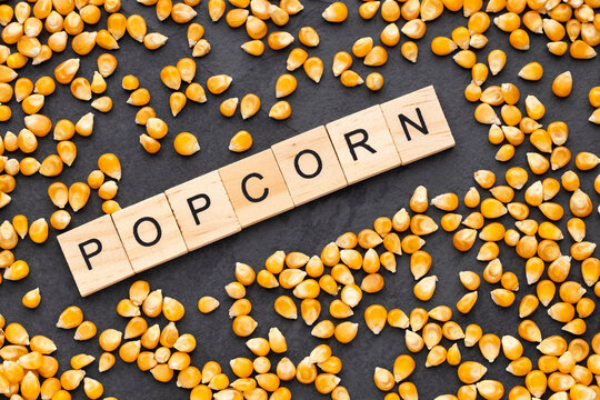 Raw yellow corn kernels scattered across a dark slate surface, accompanied by the word "POPCORN" spelled out with wooden tiles.