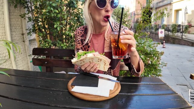 Blonde tourist sitting at a table in Brisighella, Italy, enjoying a piadina romagnola and drinking a cocktail through a straw