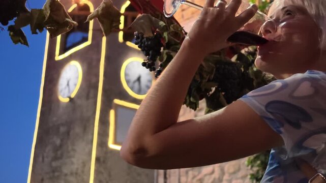 SLOW MOTION: Blonde woman sipping Lambrusco at a lively night wine festival in Castelvetro di Modena, enjoying local traditions under an illuminated historic clock tower and festive lights in Italy