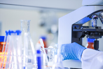 A female microbiologist in a white coat, protective eyewear and a sterile hairnet reaching past sample vials toward objective turret inside a university biotechnology laboratory. © ultramansk