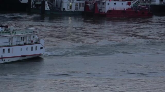 White river cruise ship moving through harbor waters at dusk, leaving a visible wake, with red and white workboats moored in background