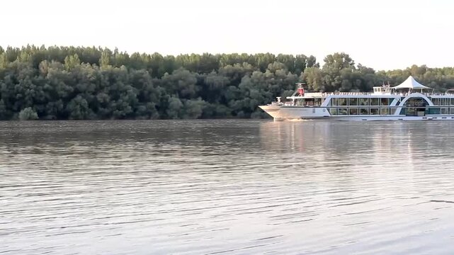 White river cruise ship STEAUA DUNARII cruising on calm water at dusk near forested shoreline with passengers on upper deck