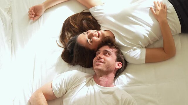 Young couple lie head to head in bed, smiling and laughing playfully in warm sunlight. Man and woman share an intimate, relaxed moment at home, expressing love, romance, partnership and togetherness.