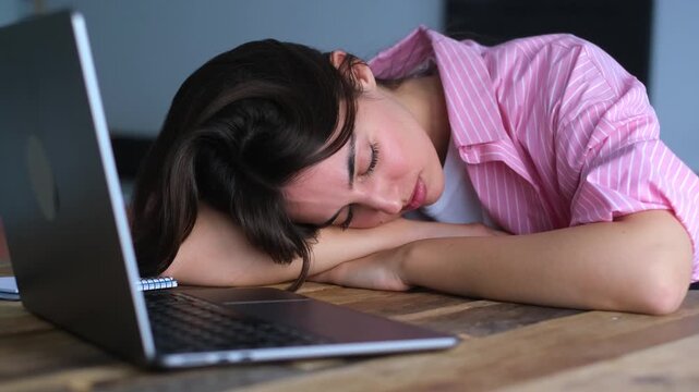 Woman sleeps beside an open laptop with her head on folded arms, showing work burnout, stress and fatigue in a home office. Close-up static clip supports workplace wellness and mental health messaging
