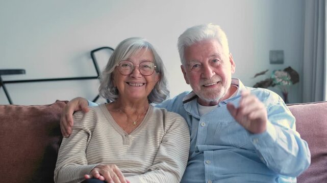 Senior couple sits on a sofa at home, smiling, waving and talking to the camera during a video call. Elderly man and woman share a cheerful, friendly remote conversation with family or close friends.