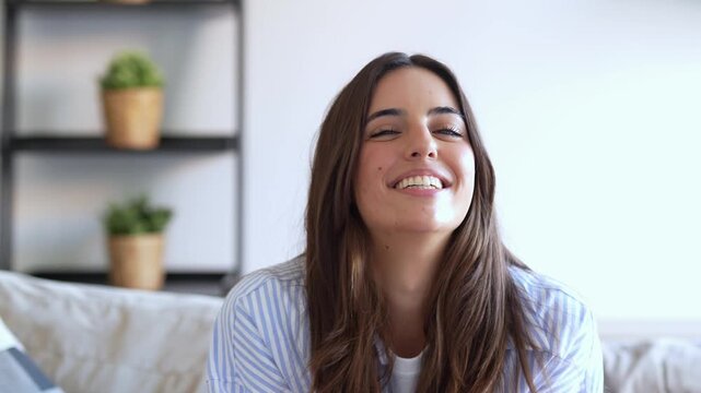 Smiling young woman nods at the camera while sitting on a sofa in a bright living room. Casual lifestyle portrait conveys happiness, agreement, warmth and a cheerful approachable mood indoors.