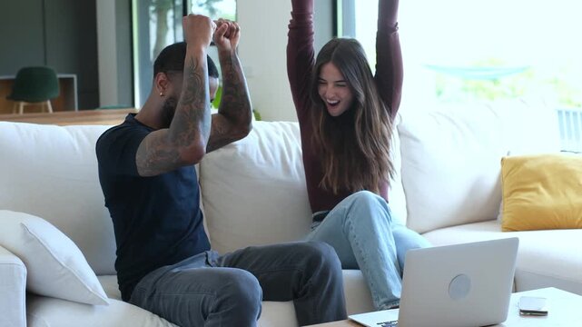 Couple watches a laptop in their bright living room, then celebrates good news with laughter, high fives and raised arms. Their joyful reaction conveys love, relationship, togetherness and success.