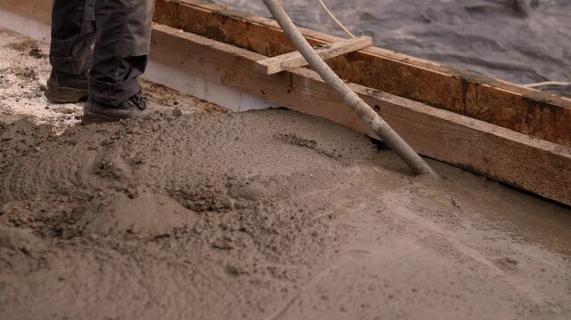 Close-up of wet concrete being leveled inside wooden formwork beside work boots and long tool, with rough muddy texture, rusted edges, warm natural light, and low angled view on active building site.