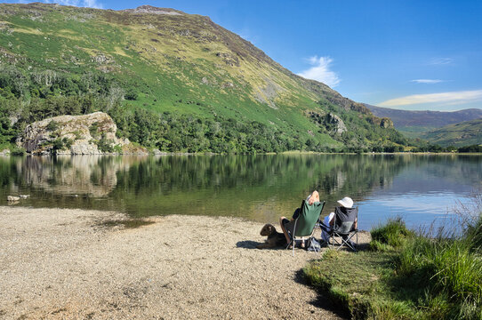 People relaxing in folding chairs on the gravel shore of Llyn Gwynant with a dog, enjoying views of the tranquil lake and surrounding mountains in Snowdonia National Park, North Wales.
