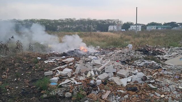 Smoldering fire and white smoke rising from open burning trash dump and scattered construction debris in an empty outdoor field, representing air pollution, illegal waste dumping, and environmental ha