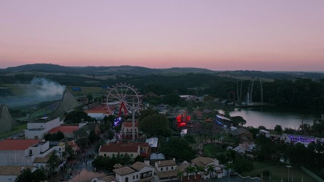 Drone pan right over Hopi Hari during Hopi Pride, showing a large LGBT event crowd, illuminated stages, Ferris wheel and amusement park at dusk.