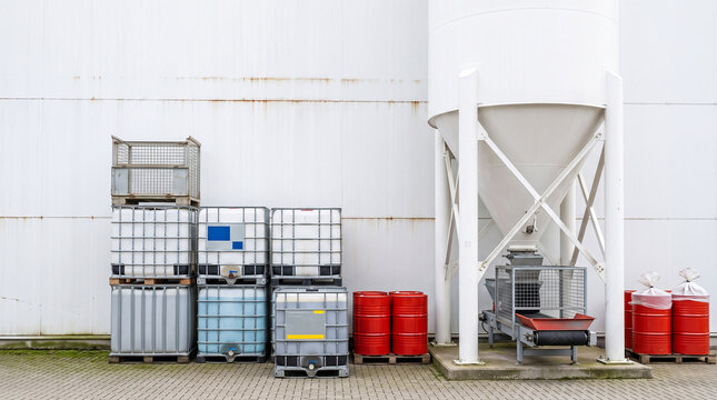 IBC totes, red steel drums, and a white industrial hopper silo at a factory exterior wall